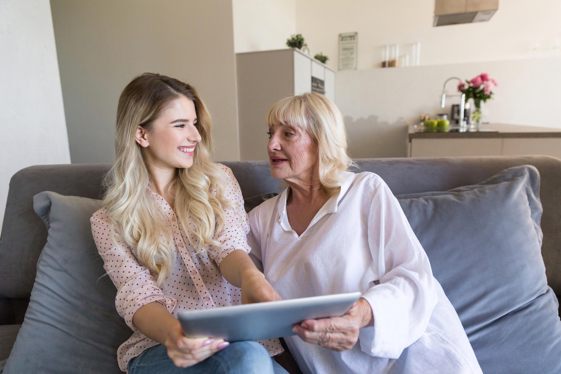 Grandmother and granddaughter sit on a couch at home while the granddaughter smiles and looks at the grandmother. They hold a large tablet together.